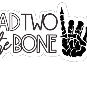 May include: A white cake topper with the phrase "BAD TWO the BONE" in black lettering, accompanied by a black skeleton hand making a rock and roll gesture. The design is outlined in black.