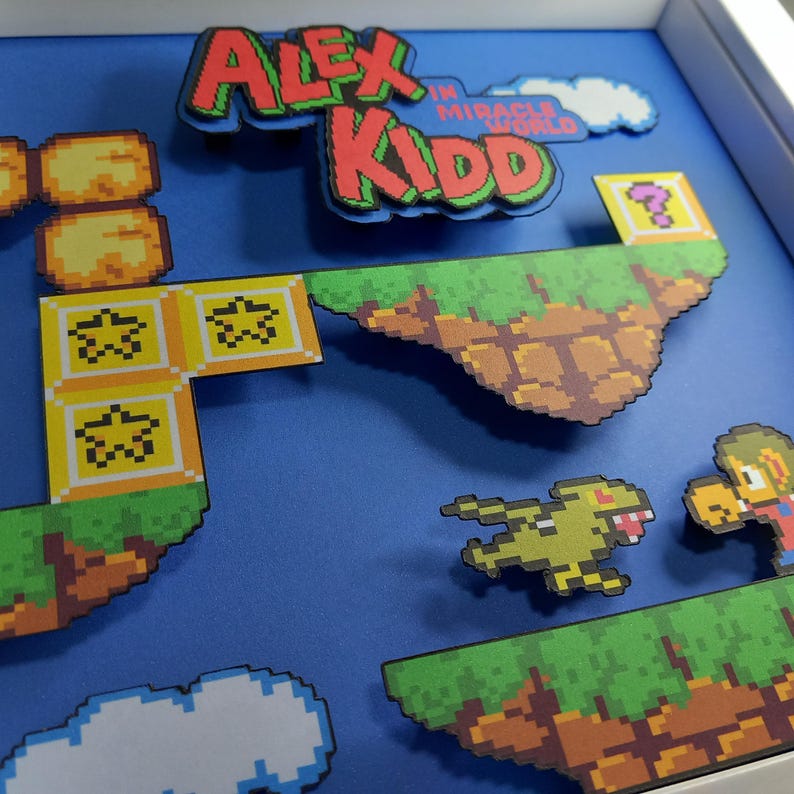 May include: A 3D papercraft diorama of the video game Alex Kidd in Miracle World. The diorama features a blue background with a green and brown platform, a yellow flying enemy, and a character from the game.