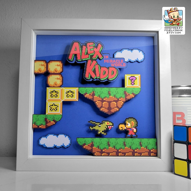 May include: A framed pixel art picture of a video game character, Alex Kidd, in a blue world with clouds, green grass, and brown rocks. The title of the game is "Alex Kidd in Miracle World".