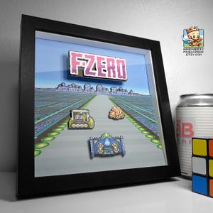 May include: A black framed shadow box with a pixelated image of a video game race track. The title "FZERO" is displayed in pink letters. Three pixelated race cars are on the track, one yellow, one orange, and one blue.