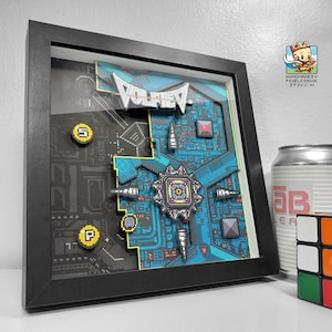 Peut inclure: Une œuvre d'art encadrée en papier superposé avec un design pixélisé dans des tons de bleu, gris et jaune. L'œuvre présente le mot "VOLFIEV" et un motif géométrique complexe. Une canette et un Rubik's Cube sont à côté du cadre.