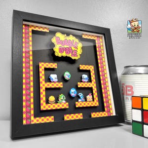 May include: A framed 3D shadow box featuring a pixelated design inspired by the classic arcade game, Bubble Bobble. The artwork includes the game's logo, characters, and level layout, with a black frame and colourful accents. The artwork is approximately 25 cm by 25 cm.