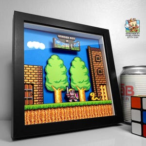 May include: Framed 3D pixel art of a classic video game scene. The artwork features a pixelated character with a sword, trees, a brick tower, and the text "Wonder Boy in Monster Land". The frame is black.