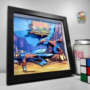 Könnte beinhalten: Gerahmtes 3D-Kunstwerk mit Charakteren aus Marvel vs. Capcom. Das Kunstwerk zeigt Chun-Li und Spider-Man in Actionposen vor einem pixeligen Hintergrund. Der Rahmen ist schwarz und das Kunstwerk wird in einem Schattenbox-Stil präsentiert.