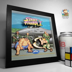 Angled view of a framed Street Fighter II pixel art shadow box showing E. Honda facing Guile on the airbase stage.