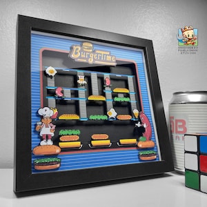 Könnte beinhalten: Gerahmtes 3D-Kunstwerk mit dem Arcade-Spiel Burgertime. Das Kunstwerk zeigt einen Kochcharakter, Burger und andere Lebensmittel. Der Rahmen ist schwarz und der Hintergrund blau mit einem Rastermuster. Eine Dose und ein Rubik's Cube sind sichtbar.