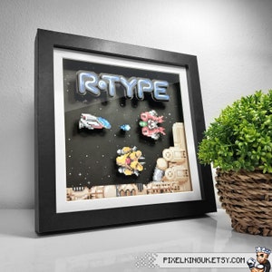 May include: A black framed shadow box featuring a pixelated image of the video game R-Type. The image includes the title "R-TYPE" and pixelated spaceships.