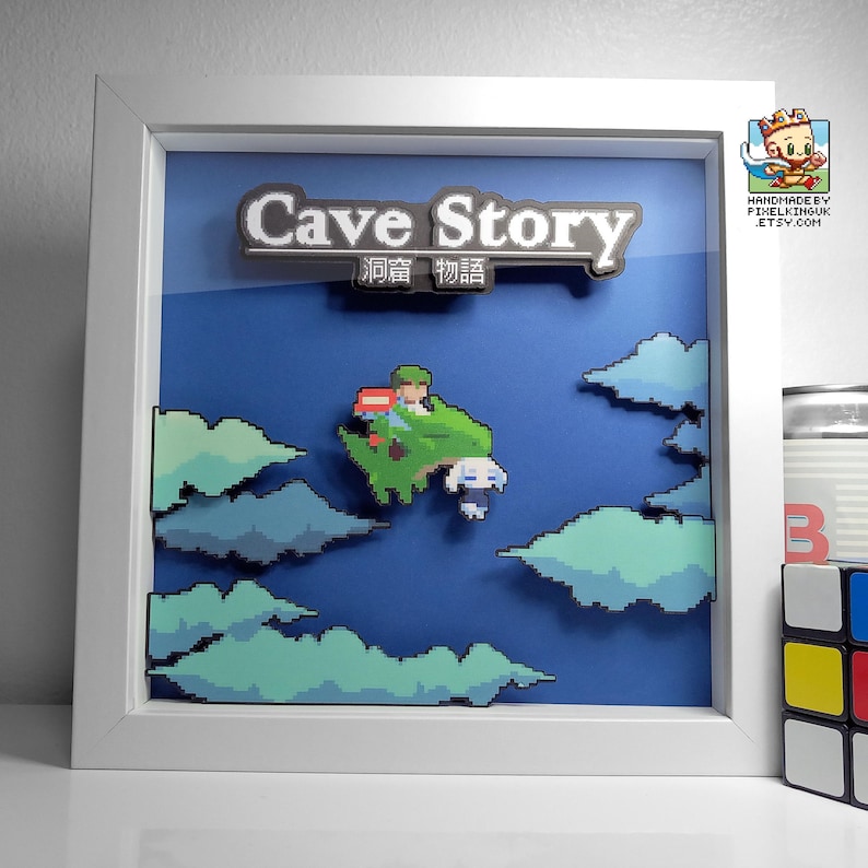 May include: A white framed shadow box with a blue background and a pixelated image of a character from the video game Cave Story. The title "Cave Story" is written in white letters on a black background. The Japanese text "洞窟物語" is written in white letters on a black background below the title.