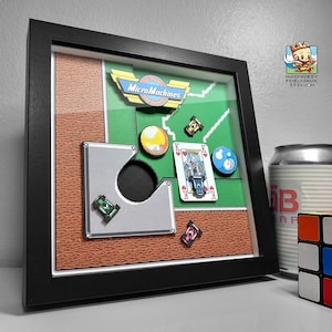 Könnte beinhalten: Gerahmtes Kunstwerk mit einem Micro Machines-Logo und Spielelementen. Es enthält eine Herz-König-Spielkarte, einen gelben Kreis, eine blaue Acht-Ball-Kugel und Miniatur-Autodesigns. Der Rahmen ist schwarz, und der Hintergrund hat grüne und braune Abschnitte.