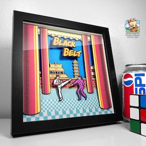 May include: Framed 3D artwork of a retro video game scene. The artwork features two pixelated figures in a martial arts fight, with the words "Black Belt" above them. The background includes a pagoda and red curtains. A can of Pepsi and a Rubik's cube are next to the frame.
