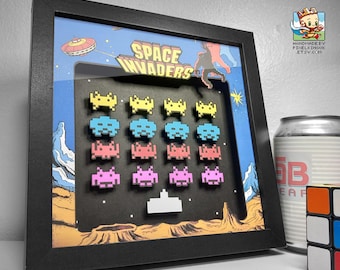 May include: A black framed shadow box with a 3D diorama of the classic arcade game Space Invaders. The diorama features colorful pixelated alien spaceships and a white pixelated spaceship at the bottom.