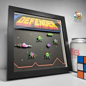 Op de afbeelding: Een zwart ingelijst beeld met een pixel art-scène van het klassieke arcade-spel Defender. De scène toont ruimteschepen, aliens en het woord "DEFENDER" in gele letters.