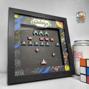 Peut inclure: Une boîte à ombre encadrée de noir avec une scène de Galaga en pixel art. La scène représente le vaisseau spatial Galaga et des ennemis extraterrestres sur un fond spatial. Le titre "Galaga" est écrit en lettres blanches sur un fond noir.