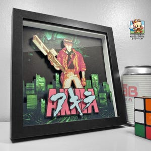 以下が含まれることがあります： アニメ「AKIRA」のキャラクターが武器を持っている様子を描いた額装アート。背景は都市景観で、ピンクと青で「AKIRA」の日本語タイトルが描かれています。黒いフレームに入っています。
