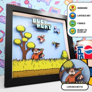 Może przedstawiać: Czarna ramka typu shadow box z warstwową grafiką pikselową z gry wideo Duck Hunt. Obraz przedstawia brązowego psa trzymającego kaczkę, drzewo, trawę i latające kaczki na jasnoniebieskim tle z napisem DUCK HUNT.