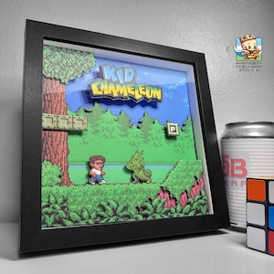 May include: A framed pixel art piece of the video game Kid Chameleon. The art is in a black frame and features a green background with a character in blue and a green creature. The title "Kid Chameleon" is in the upper center of the image.