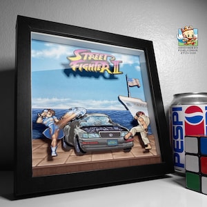 Puede incluir: Una obra de arte 3D enmarcada que representa una escena de Street Fighter II con personajes, un coche y un barco contra un fondo de cielo azul. La obra incluye el logotipo del juego y está enmarcada en negro. Una lata de Pepsi y un Cubo de Rubik están al lado.