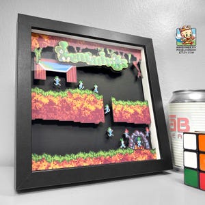 May include: A framed 3D pixel art scene depicting a video game level with green, red, and brown pixelated terrain. The artwork includes the word "Lemmings" and small pixelated characters. The frame is black.