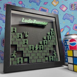 May include: A framed 3D artwork featuring the text "LodeRunner" in green, with a pixelated landscape design. The artwork is in a black frame, with a can of Pepsi and a Rubik's Cube next to it.
