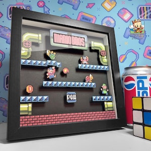 May include: A framed 3D shadow box featuring a pixelated scene from the video game Mario Bros. The artwork includes characters, platforms, and the text "MARIO BROS." in a black frame. A can of Pepsi and a Rubik's cube are in the background.