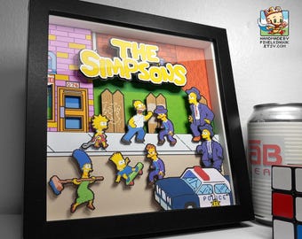 Puede incluir: Obra de arte 3D enmarcada con los personajes de Los Simpson y el logo del programa. La escena incluye personajes como Homer, Bart y Marge, con un coche de policía y un fondo colorido. El marco es negro.