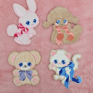 Puede incluir: Cuatro parches bordados para planchar que representan un conejito, un caniche, un osito de peluche y un gatito. Cada animal lleva un lazo y tiene ojos azules.