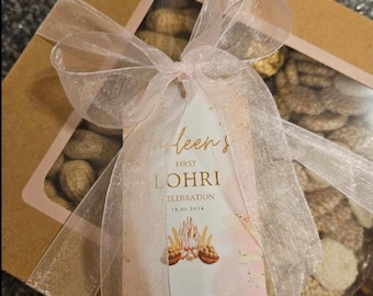 May include: A brown gift box with a pink ribbon and a tag that says "First Lohri Celebration 13.01.2023". The box is filled with a variety of snacks, including peanuts, puffed rice, and other treats.