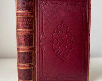 May include: A vintage book with a deep red leather cover and gold accents. The spine features the title "Habbey's Poetical Works" in gold lettering. The front cover has an embossed floral design. The book's edges are also trimmed in gold.