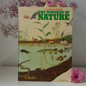 以下が含まれることがあります： 「Looking Inside The Wonders of Nature」というタイトルのヴィンテージ本。魚や鳥など、野生生物のイラストが描かれています。本はベージュ色の表紙で、緑と青の文字とイラストが描かれています。ピンクのバラとピンクの花が背景にあります。