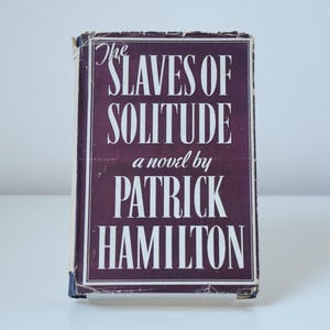 May include: A burgundy book cover with white text that reads "The Slaves of Solitude, a novel by Patrick Hamilton".