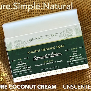 May include: A white box with green accents and a label that reads "HEART TONE ANCIENT ORGANIC SOAP Coconut Créme" with the text "Made with ORGANIC COCONUT MILK NET WT 5 Oz/150g GMO FREE ALL NATURAL USES FACE BODY MOISTURIZING SENSITIVE SKIN RICH SHAVING LATHER VEGAN PLANT BASED" on the box. The box is sitting on a brown hessian surface. The text "PURE COCONUT CREAM UNSCENTED" is below the box.