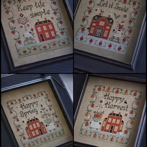 Peut inclure: Quatre broderies au point de croix encadrées sur des thèmes saisonniers. Chacune présente une maison rouge, des bordures florales et du texte : « Keep life simple », « Let it snow », « Happy Spring » et « Happy Harvest ». Les cadres sont en bois foncé.
