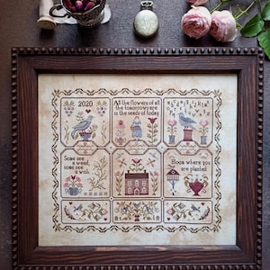 Può includere: Un sampler a punto croce incorniciato con una cornice in legno marrone scuro. Il sampler presenta motivi floreali e di uccelli, con testo tra cui "2020" e "Bloom where you are planted". La tavolozza dei colori include sfumature di marrone, rosa e blu.