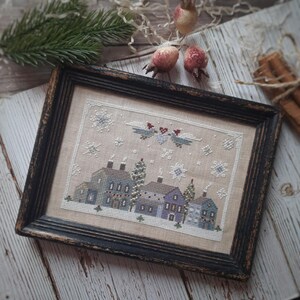 May include: Cross-stitch artwork in a distressed black wooden frame. The artwork depicts a winter village scene with houses in shades of blue and purple, snowflakes, and a pair of birds. The background is a neutral beige color.