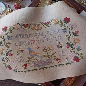 Puede incluir: Un muestrario de punto de cruz beige con un borde floral y el alfabeto. El diseño incluye un pájaro azul, flores en rojo, amarillo y rosa, y los números 20 y 21. El muestrario está sobre un trozo de tela de lino.