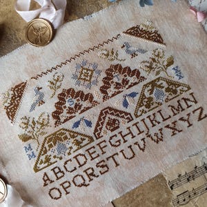 May include: A cross-stitch sampler with a beige background features brown, blue, and green floral and geometric patterns. The alphabet is stitched in brown. The sampler has a vintage aesthetic, with a torn edge and a handwritten note.