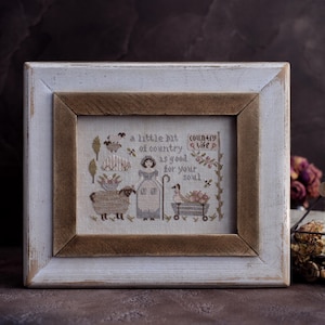 Puede incluir: Un bordado de punto de cruz enmarcado con un tema rústico y campestre. La obra de arte presenta una oveja, una mujer y un pato con un carro, con el texto "a little bit of country life is good for your soul". El marco es blanco con un borde interior marrón.