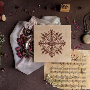Può includere: Un tessuto quadrato beige con un disegno floreale a punto croce marrone è il punto focale. L'immagine include un orologio da taschino vintage, boccioli di rosa secchi, spartiti musicali e un timbro di legno con una rosa.