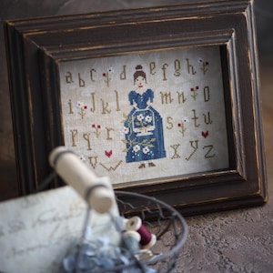 Könnte beinhalten: Ein gerahmtes Kreuzstich-Exemplar mit einer Frau in einem blauen Kleid, umgeben von einem Alphabet und floralen Akzenten. Der Rahmen ist dunkelbraun. Ein Drahtkorb mit Garnrollen und Papier befindet sich im Vordergrund.