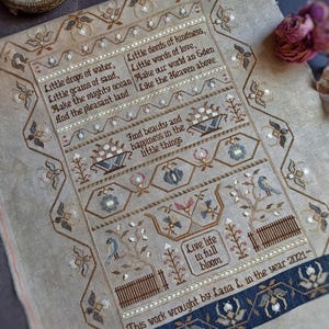 May include: A detailed cross-stitch sampler with inspirational quotes and floral motifs. The piece features text such as "Little drops of water" and "Live life in full bloom." The color palette includes beige, brown, blue, and white threads, creating a vintage aesthetic.