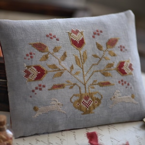 Peut inclure: Un coussin carré gris clair avec un motif floral brodé au point de croix. Le motif présente des fleurs rouges et dorées dans un vase, avec deux petits lapins. Le coussin est probablement fait à la main et a une esthétique rustique et vintage.