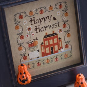 Puede incluir: Obra de arte de punto de cruz en un marco negro con un diseño de "Happy Harvest". La obra representa una casa roja, calabazas y una oveja. Dos decoraciones de calabaza naranja están en primer plano. La obra está enmarcada en un marco de madera negro.