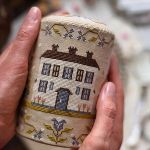 May include: A cylindrical, hand-stitched needlework container featuring a house design. The beige fabric is adorned with a brown roof, blue door, and floral accents. The container is held in two hands.