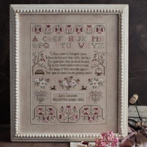 Op de afbeelding: Een ingelijste kruissteeksampler met een crèmekleurige achtergrond. De sampler heeft bloemen en geometrische patronen in tinten bordeauxrood en groen, met het alfabet en een gedicht. De lijst is wit met een parelrand.
