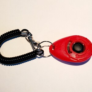 May include: A red dog training clicker with a black button and a black coiled wrist strap. The clicker is oval-shaped and has a metal clip and ring for attachment. The wrist strap is made of a flexible, springy material.