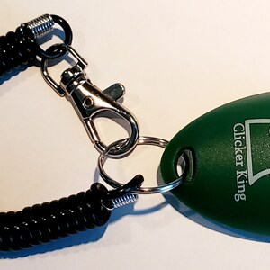 May include: A green, egg-shaped pet training clicker with the text "Clicker King" and a dog silhouette. It is attached to a black coiled wrist strap with a metal clasp and key ring.
