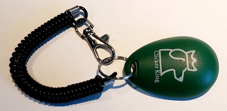 May include: A green, egg-shaped pet training clicker with the text "Clicker King" and a dog silhouette. It is attached to a black coiled wrist strap with a metal clasp and key ring.