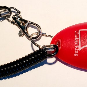 May include: A red, egg-shaped dog training clicker with the text "Clicker King" and a white graphic. It has a black coiled wrist strap and a silver-coloured metal clasp and ring.