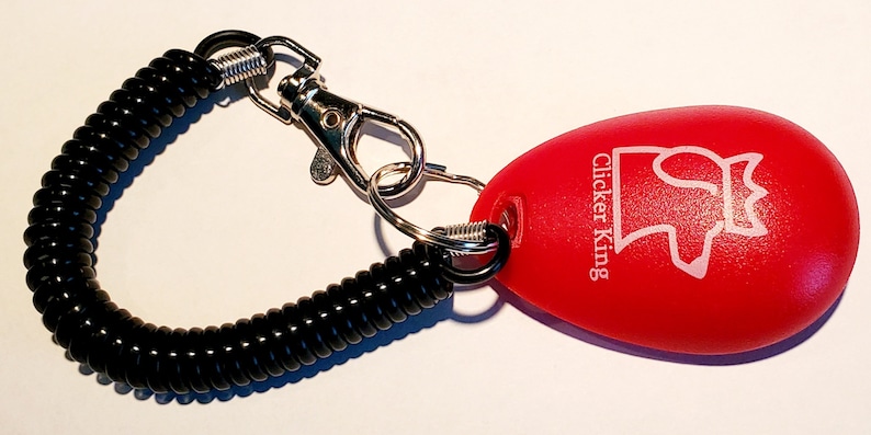 May include: A red, egg-shaped dog training clicker with the text "Clicker King" and a white graphic. It has a black coiled wrist strap and a silver-coloured metal clasp and ring.
