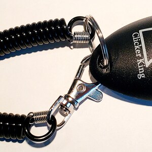 May include: Black clicker with a coiled black cord and a silver clasp. The clicker has the words "Clicker King" and a crown logo. This training tool is designed for pet training.